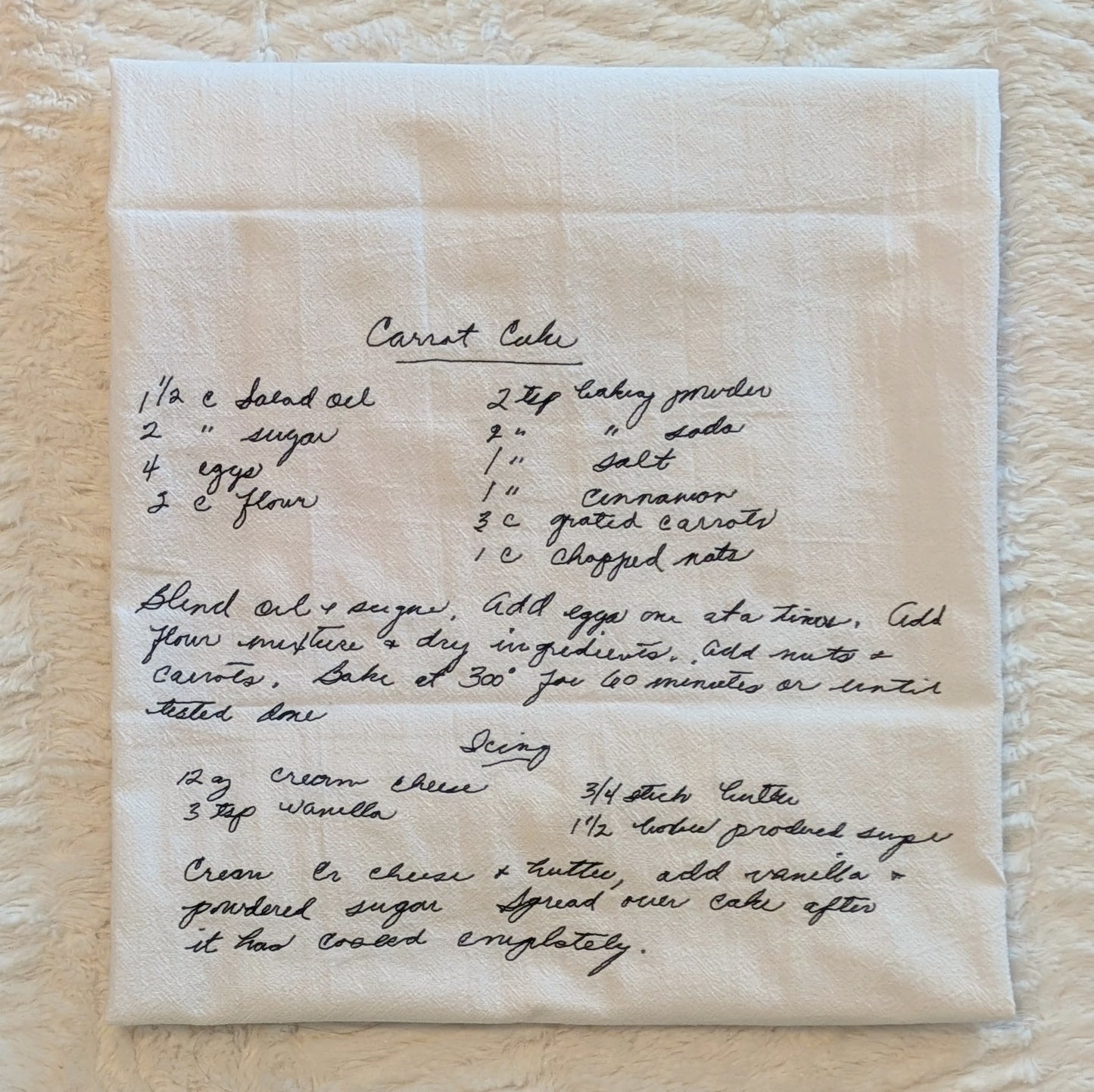 An off-white linen kitchen towel with a handwritten recipe for Carrot Cake printed in black ink, laid flat on a textured cream fabric background.