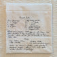 An off-white linen kitchen towel with a handwritten recipe for Carrot Cake printed in black ink, laid flat on a textured cream fabric background.